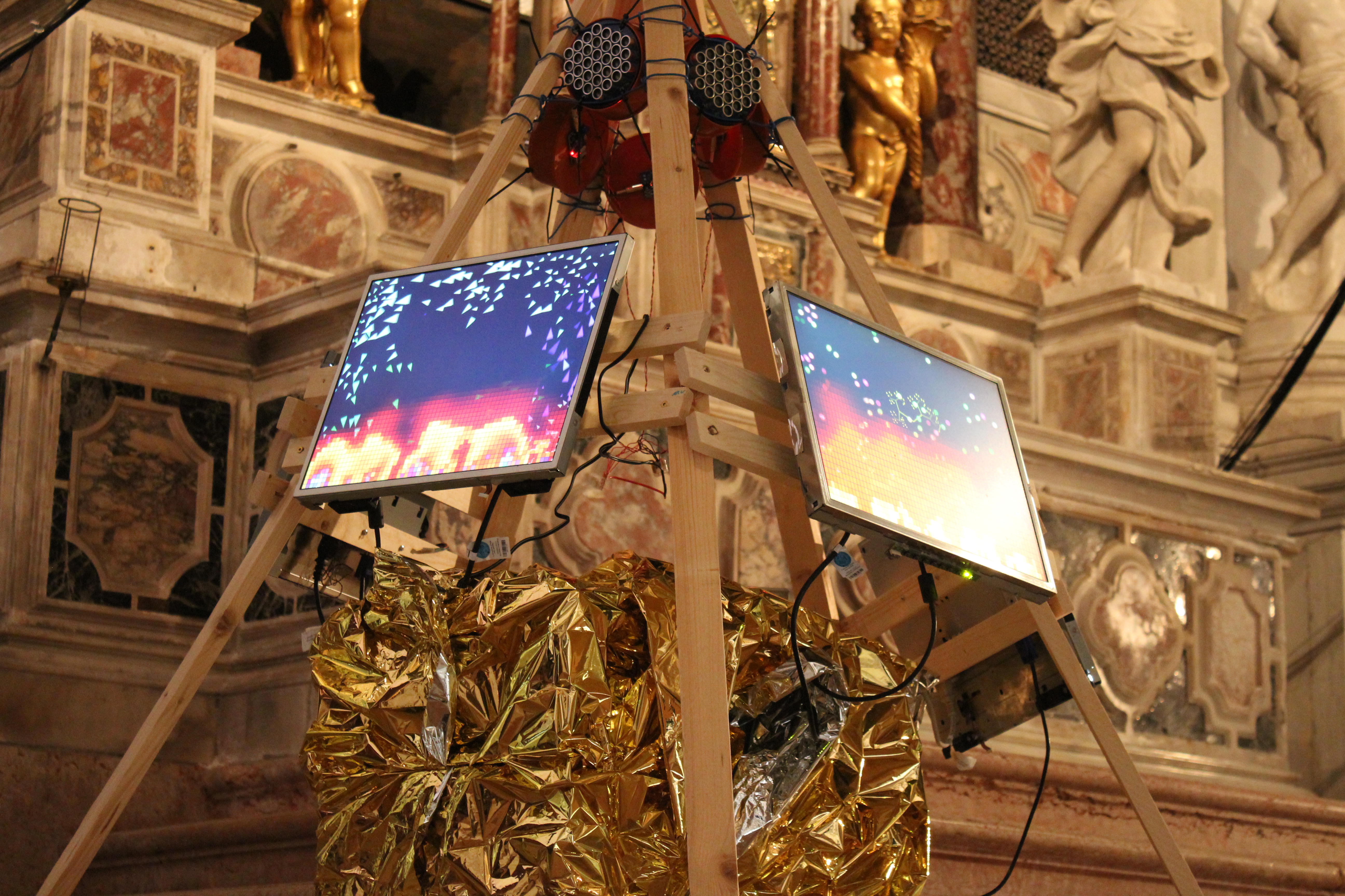 a photo of a wooden installation, pyramid-shaped arranged wooden planks holding up moitors showing processing animations, the bottom covered in golden foil, standing inside a venetian church. An altar can be seen in the background.